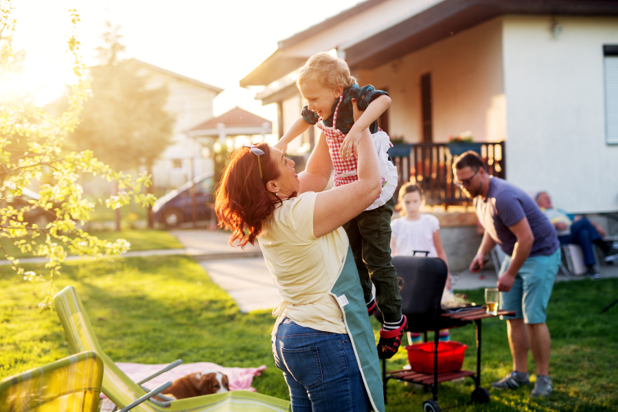 Woman is lifting up her cheerful toddler boy while the rest of her family enjoys grilling food together in their back yard